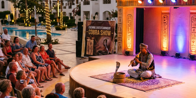Musician playing flute to a cobra on stage with audience watching outdoors at sunset